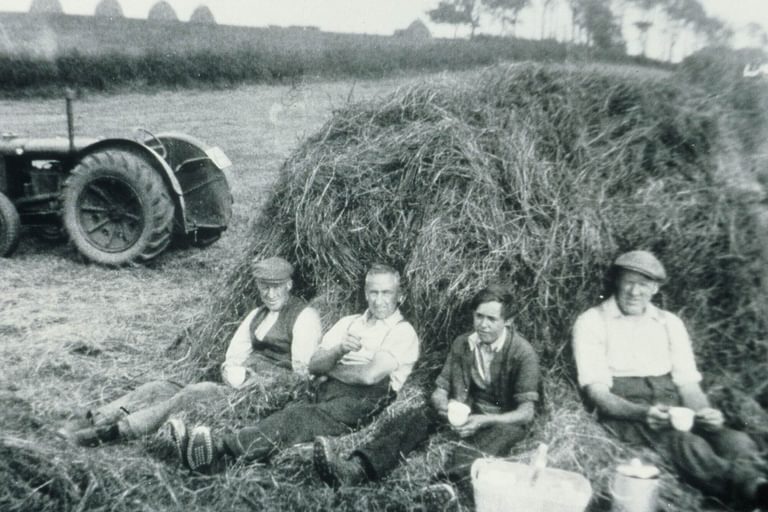 A black and white photograph of four farmers sitting against a haybale with cups in their hands on a farm.