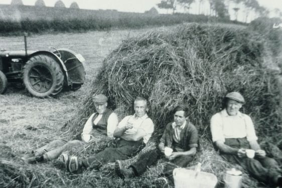 A black and white photograph of four farmers sitting against a haybale with cups in their hands on a farm.