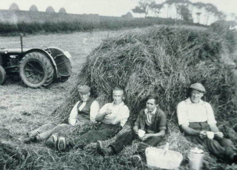 A black and white photograph of four farmers sitting against a haybale with cups in their hands on a farm.