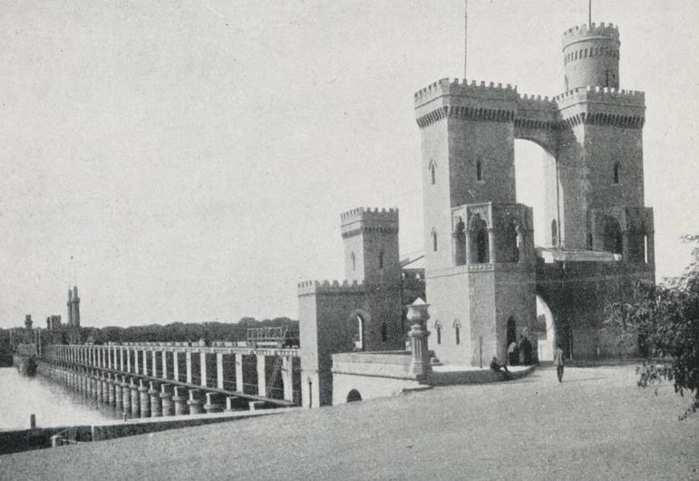 A black and white photograph of a building with tall towers alongside a river.