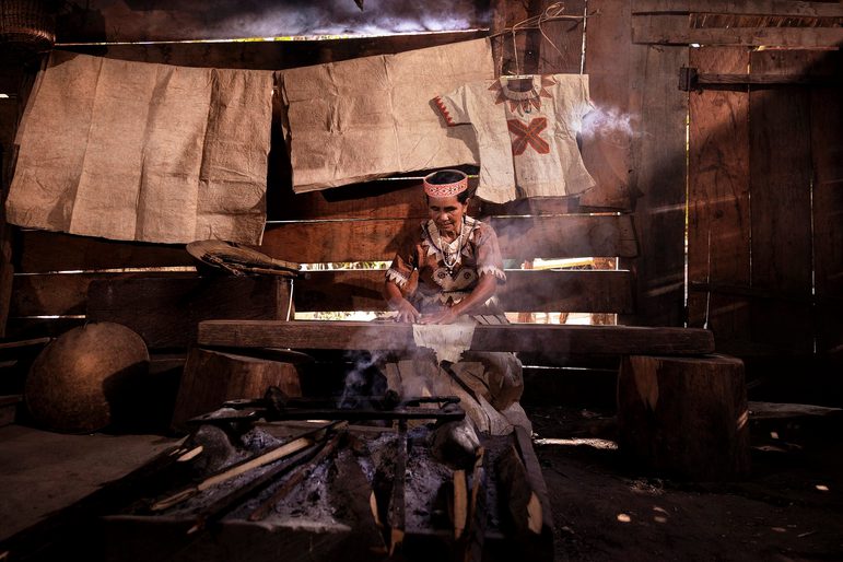 A woman sits in a dark wooded room working with textiles at a wooden bench. There is a smoldering fire in the foreground emitting smoke, and plain textiles hanging up behind her.