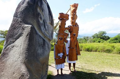 Two people in white and orange costumes stand beside a large rock with grass and trees in the background.
