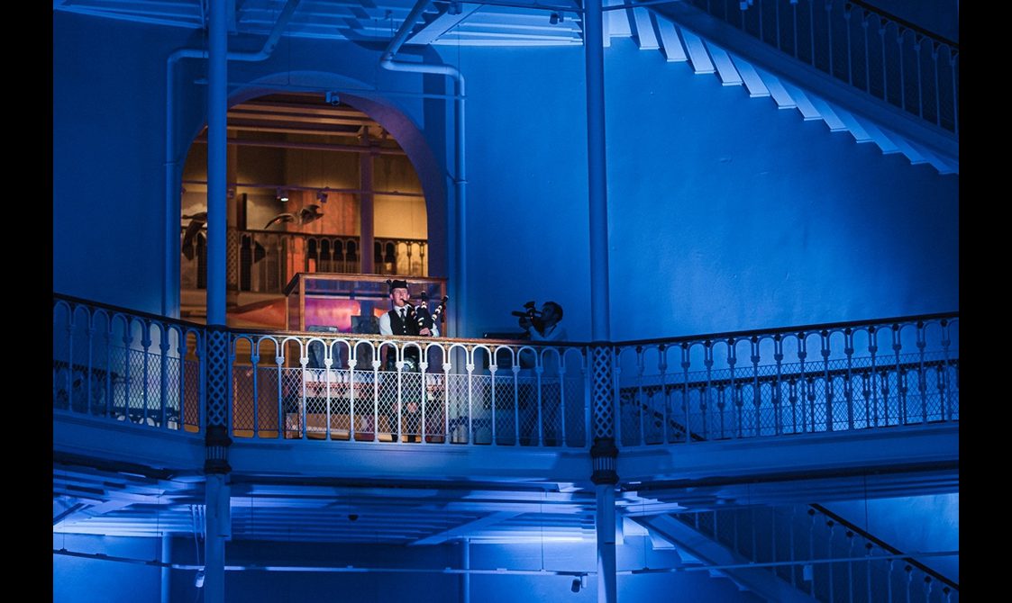 A piper playing on a balcony of a multi-level museum gallery. The space is illuminated with blue light and there is a spotlight on the piper.