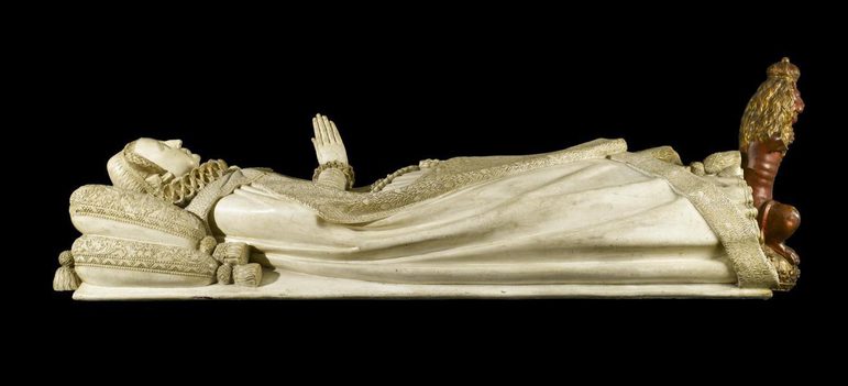 A cream sculpture of a woman wearing a long cloak and a ruffled collar lying down with cushions at her head. She has her hands held together in prayer and is posed as if on her deathbed.