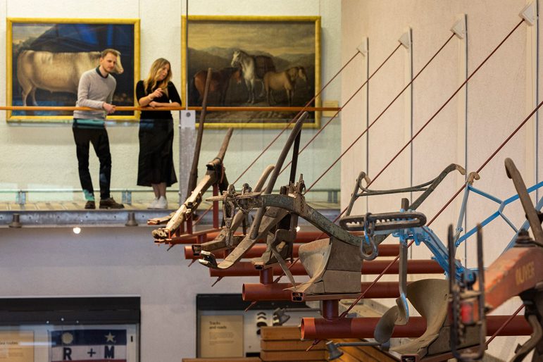 2 people are standing on a higher level in a museum, looking down at a wall full of ploughs on display.