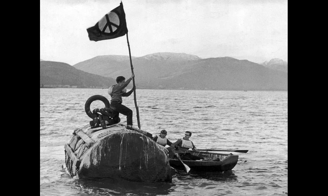 A black and white photo of a person holding a flag, standing on a submarine which is at the surface of a loch.