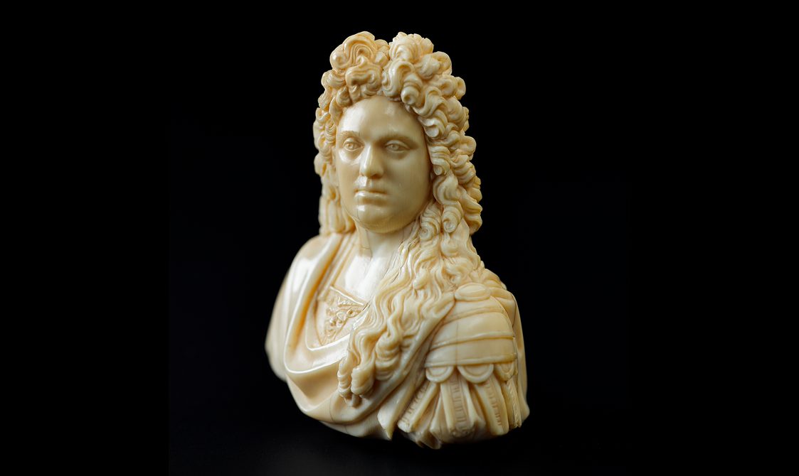 A small ivory statue of a man from the shoulders up. He has long curly hair and is wearing an elaborate draped shirt