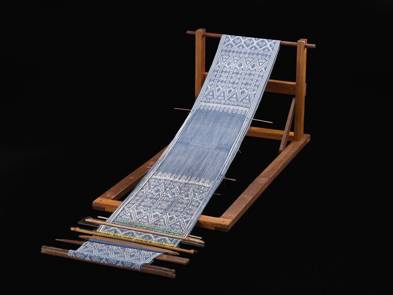 Long, narrow blue cloth with intricate white patterns in progress on a dark wooden loom.