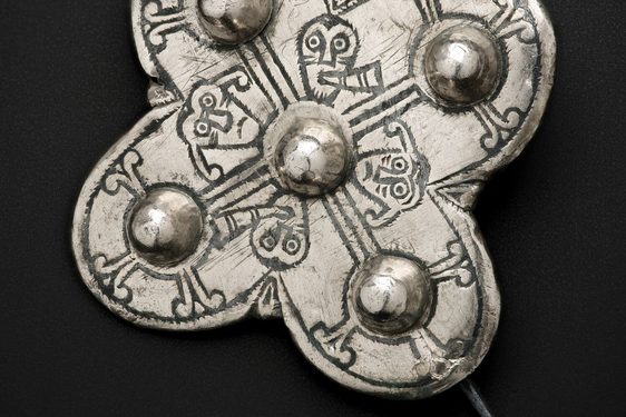 A silver brooch with a pin fastening. It is in a cross shape with a ball protruding from each point and faces and patterns inscribed on its surface.