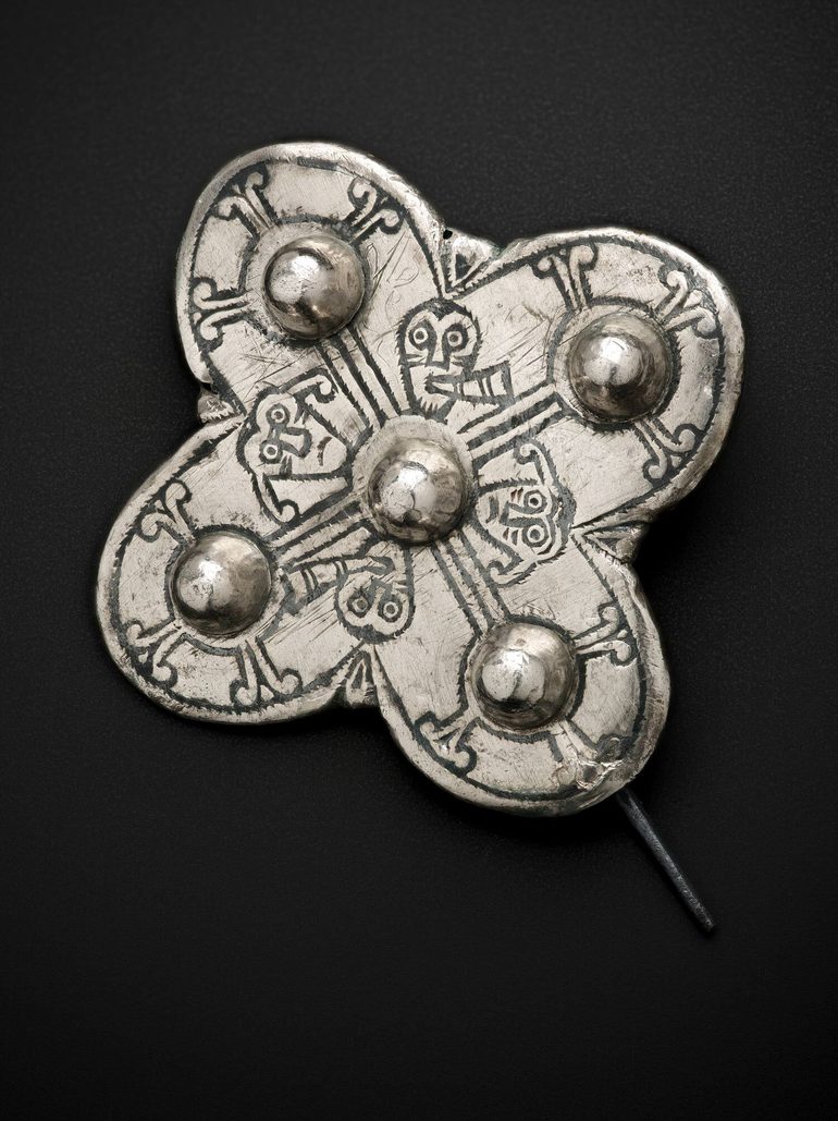 A silver brooch with a pin fastening. It is in a cross shape with a ball protruding from each point and faces and patterns inscribed on its surface.