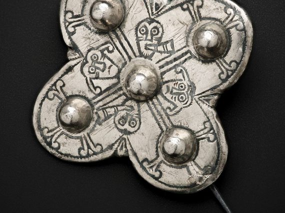 A silver brooch with a pin fastening. It is in a cross shape with a ball protruding from each point and faces and patterns inscribed on its surface.