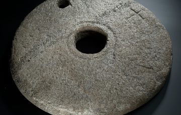 A round stone with a hole in the middle and a smaller hole to one side. There are faint carvings on the stone.