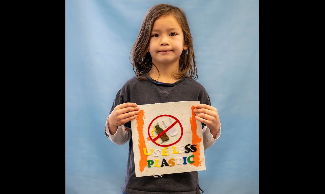 A young child is holding up a quilted square, with a note written on it that says 'use less plastic'