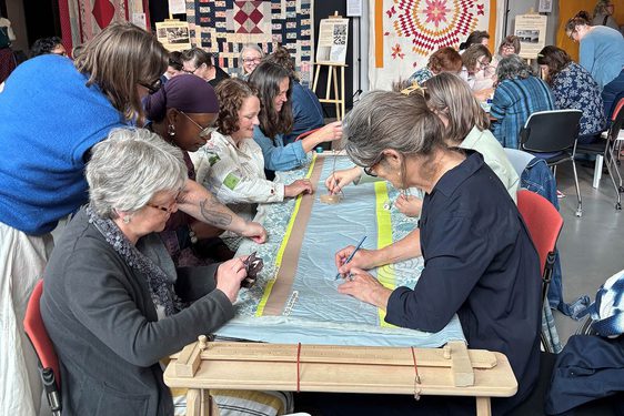 A group of people sit at a table quilting fabric.
