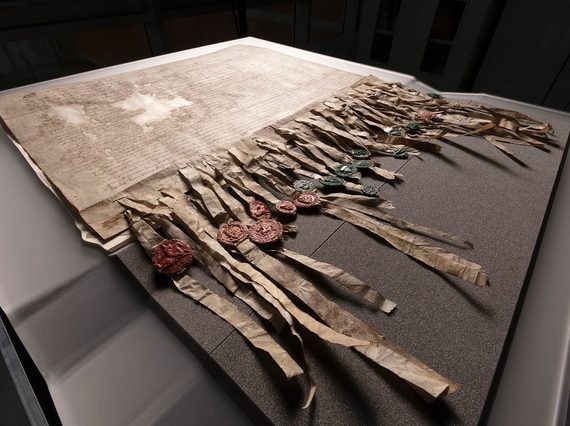 The Declaration of Arbroath laid out on a grey foam protective base set against a black background. The Declaration has dense Latin script across it with two small patches missing, and many strips dangling from the bottom affixed with green and red wax seals.