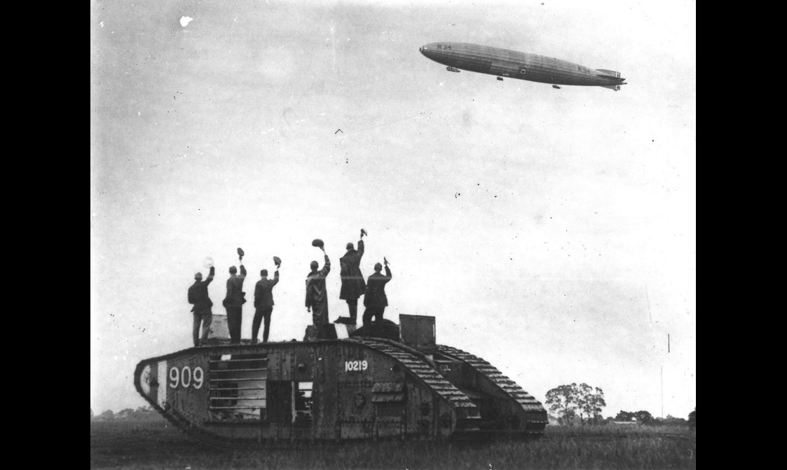 A black and white photograph of men standing on an army tank waving their hats to a blimp like airship in the sky.