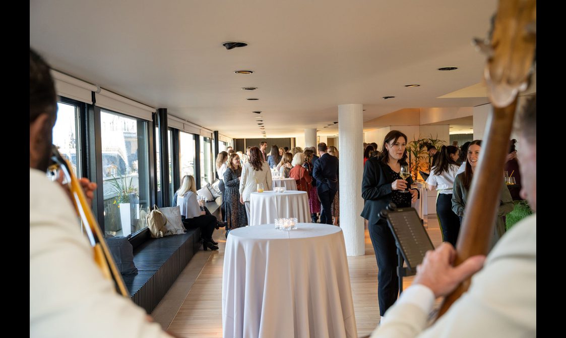 A busy event space set out for a drinks reception. People are drinking and chatting while a string quartet plays music.