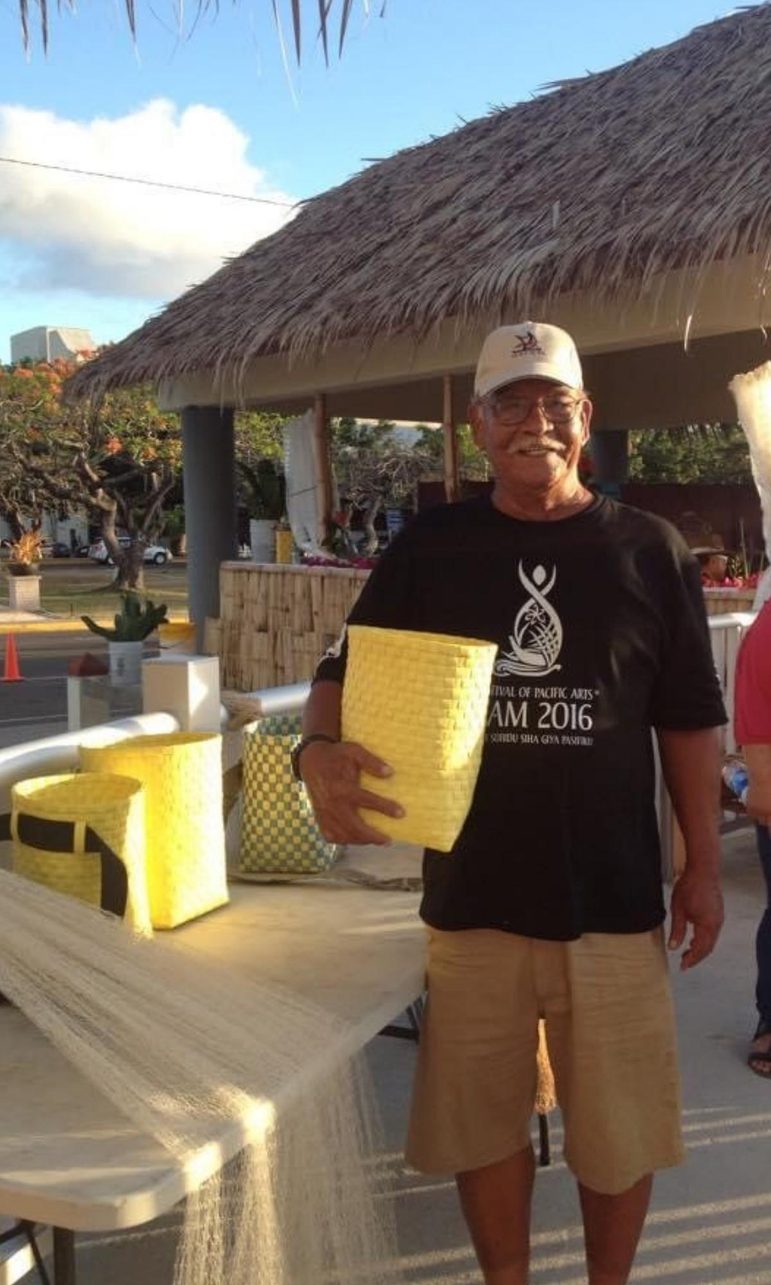 A man with glasses and a mustache, wearing a white baseball hat, black tshirt, and tan shorts, stands outside near an open structure with a grass roof. He is holding a yellow basket woven out of plastic.