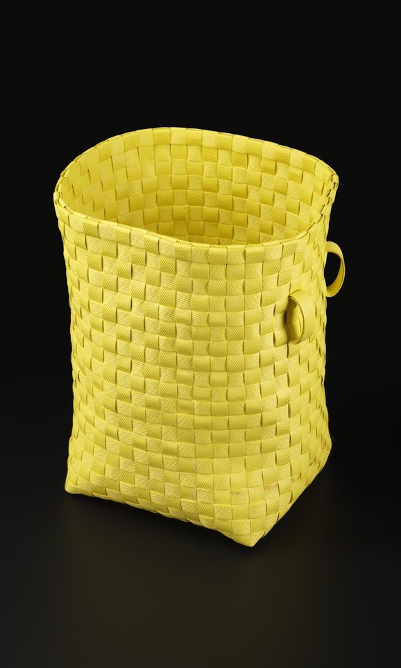 A tall, square yellow basket woven out of strips of plastic. There are two small loops near the top of one side.