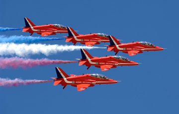 Five red arrow aircrafts flying in a blue sky in formation with red, white, and blue plumes