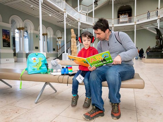 A man and a boy sit on a bench in the grand gallery looking at a large dinosaur picture book. The boy is wearing ear protectors.