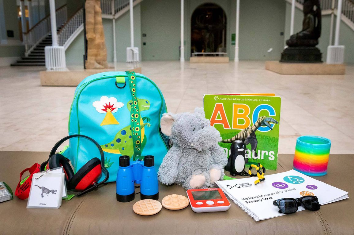 A selection of sensory objects on a bench in a museum gallery. Some of the objects include ear defenders, a cuddly elephant toy, binoculars, and sunglasses.