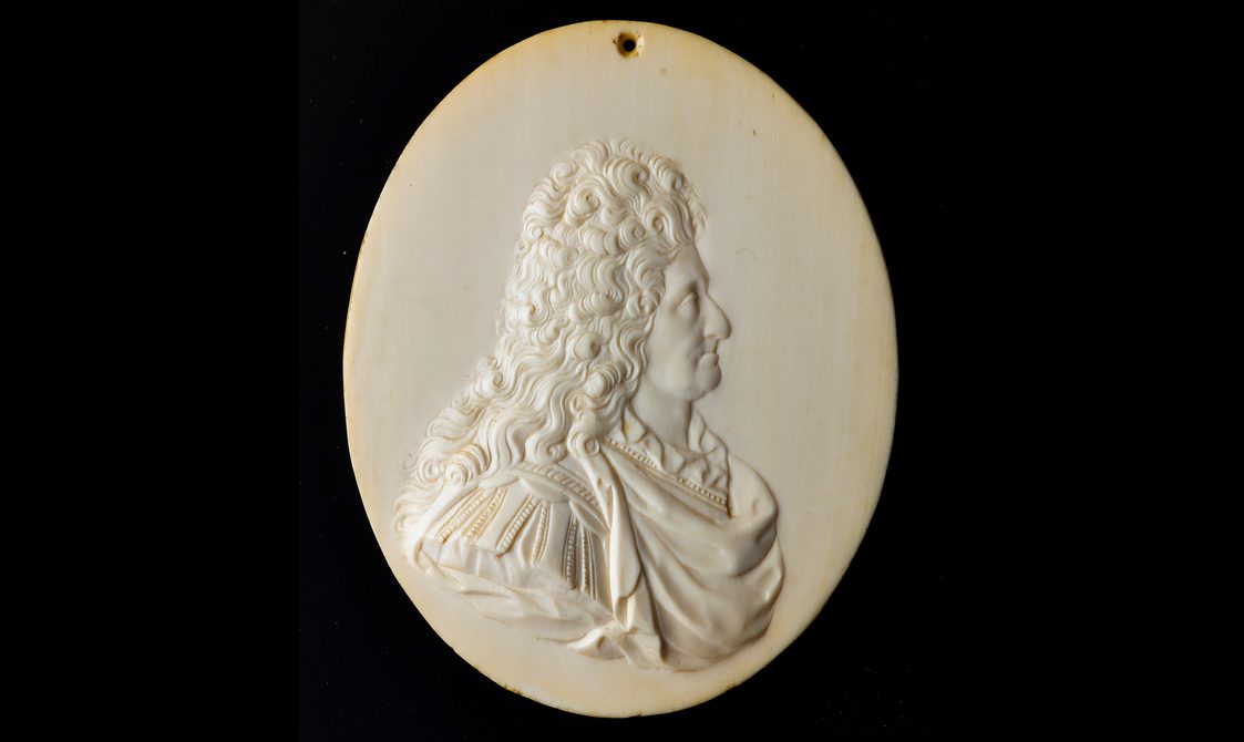 An oval ivory object with a side profile of a man carved in relief. The man has long curly hair and is wearing an elaborate draped shirt and there is text underneath the figure
