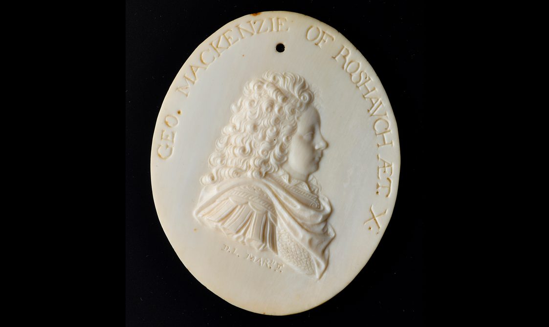 An oval ivory object with a man carved in relief. The man has long curly hair and is wearing an elaborate draped shirt and there is text carved around the edge.