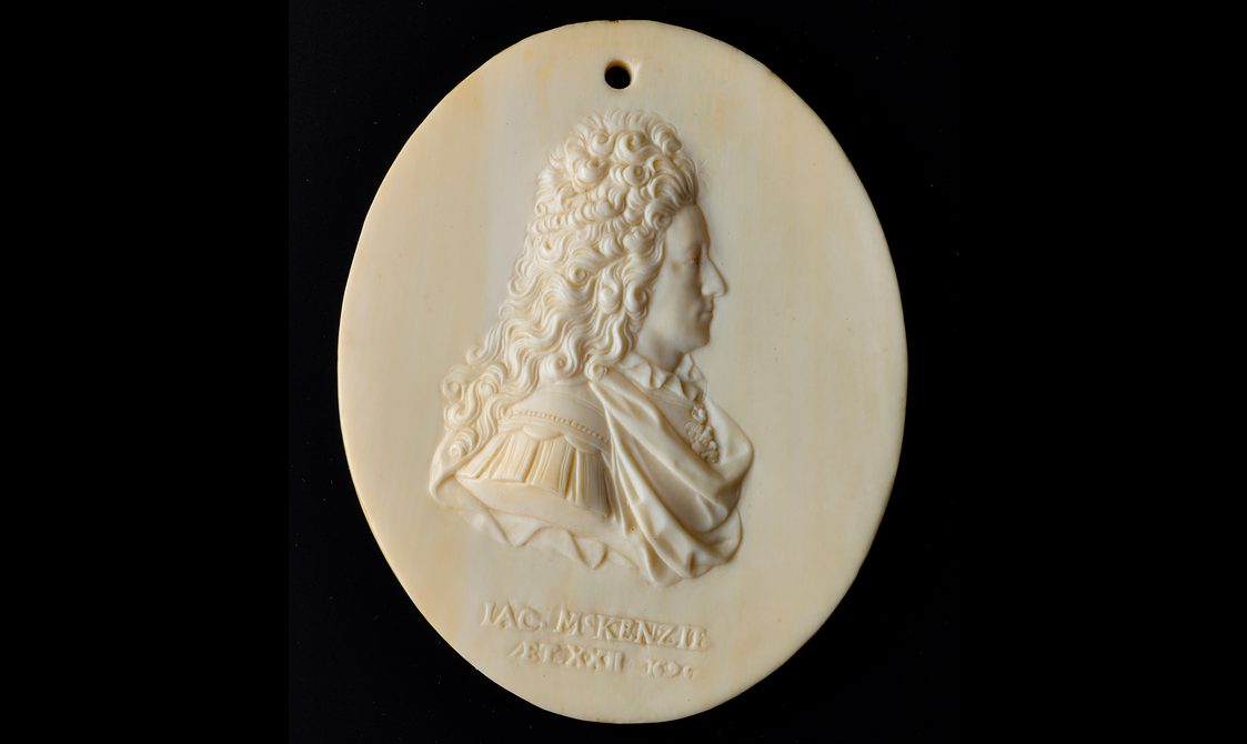 An oval ivory object with a side profile of a man carved in relief. The man has long curly hair and is wearing an elaborate draped shirt and there is text underneath the figure