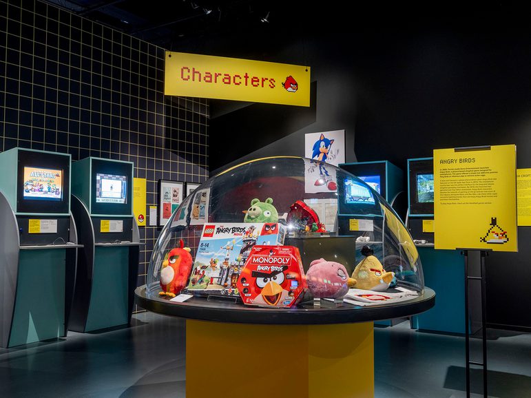 The Game On exhibition, showing consoles in the background, and a domed glass case with toys and characters from video games underneath.