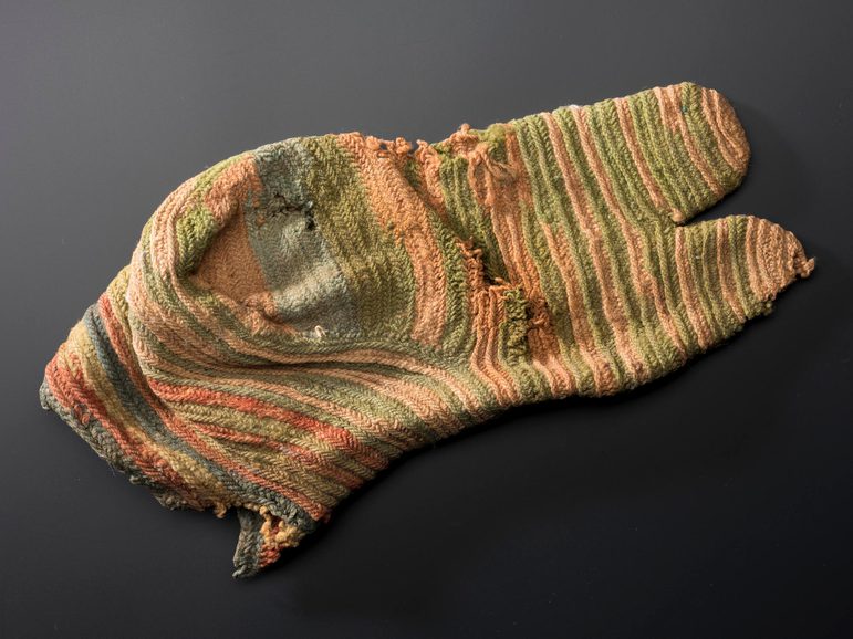The underside of a faded striped knitted sock with a split toe design and various holes.