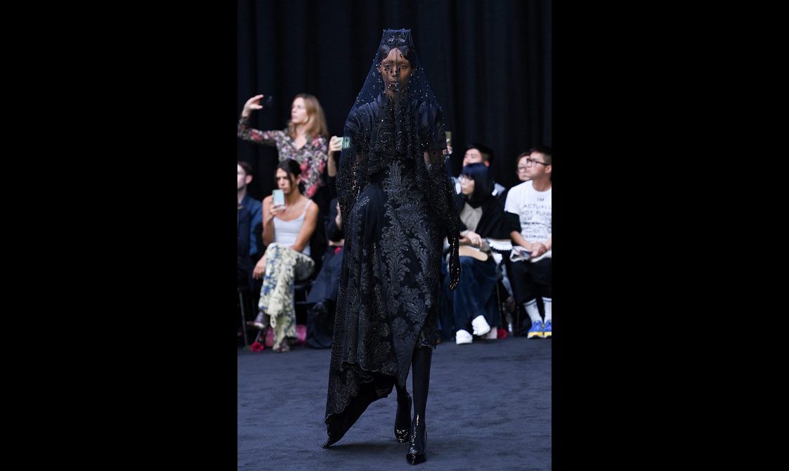 A model wearing a long black dress with black leather boots and an elaborate black veil.