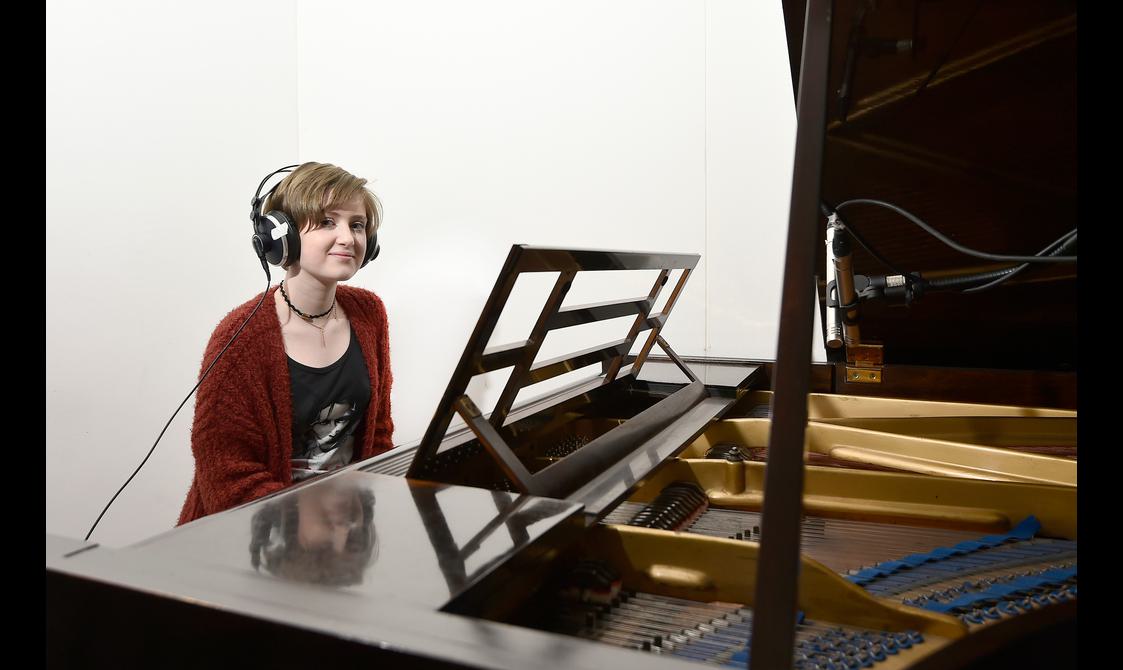A girl with short hair and a red cardigan wears large, over-ear headphones and sits at a partially-visible grand piano with the top open.