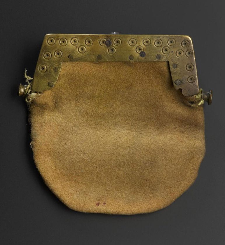 A close up of a yellow purse in a shape of a circle, with upside down U shaped brass attached to it. The brass is incised with dot-and-circle ornament.