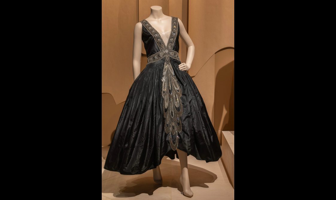 A mannequin wearing a black dress with gold detailed band around the neck and waistline