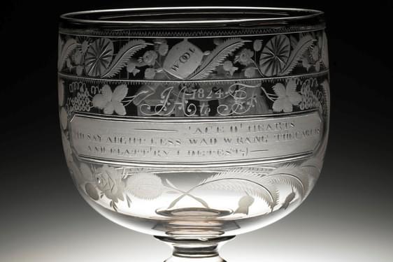 A glass goblet with plants and flowers engraved on the cup, as well as a quote. The stem is short and it sits on a small glass square base.