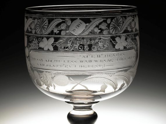 A glass goblet with plants and flowers engraved on the cup, as well as a quote. The stem is short and it sits on a small glass square base.