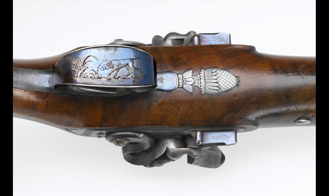 A close up of the bottom metal trigger guard, showing an engraving of a dog in a pond with long grass, and a Scottish thistle engraving attached to the guard.