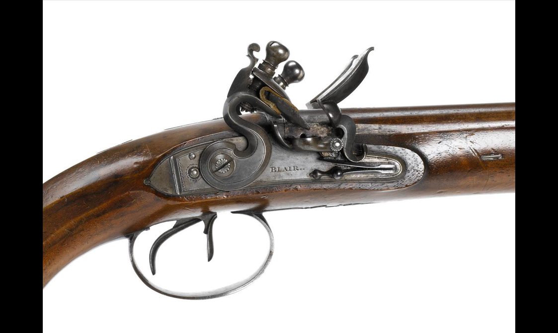 A close up detail of a wood and metal pistol showing the mechanism detail, inscribed with "Blair" to the side.