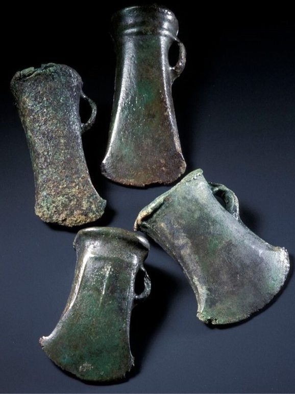 Four axe heads on a dark blue surface, tinted green with the blades facing toward the viewer.