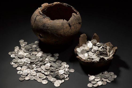 On left, a large, empty dark brown pot missing its lid with a pile of silver coins in front of it. On right, the basin of another container filled with silver coins.