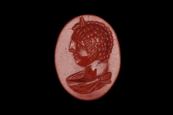 Small red gemstone that looks almost like a wax seal. Egg-shaped, with a male figure sporting right curls and large nose in profile facing left.