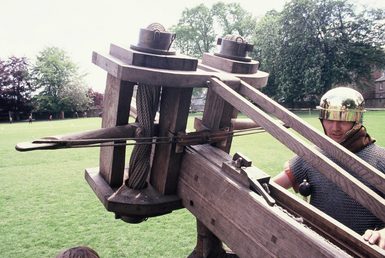A re-enactor dressed as a Roman legionary prepares a large crossbow-like ballista to fire across an open field.