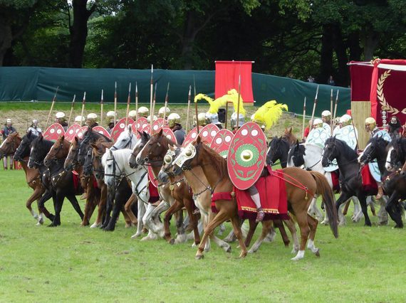 A group of people dressed like Roman soldiers on horses charge forward in a field