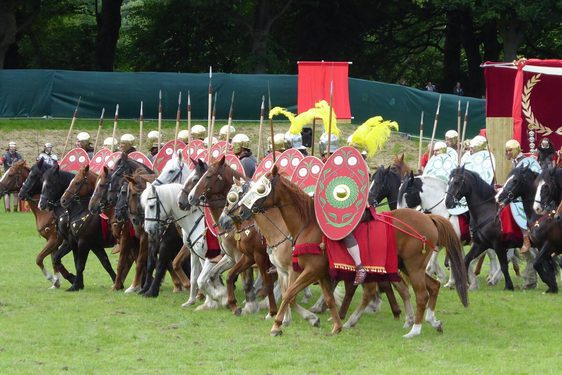 A group of people dressed like Roman soldiers on horses charge forward in a field