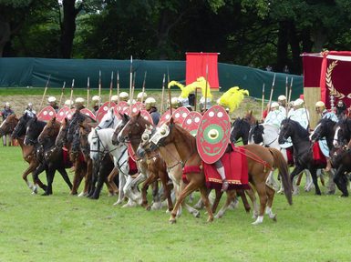 A group of people dressed like Roman soldiers on horses charge forward in a field
