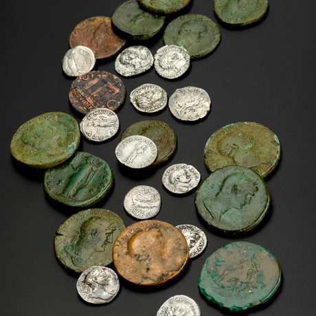 Two dozen coins spread on a dark surface, ranging from large and rusted green to small and shiny silver.
