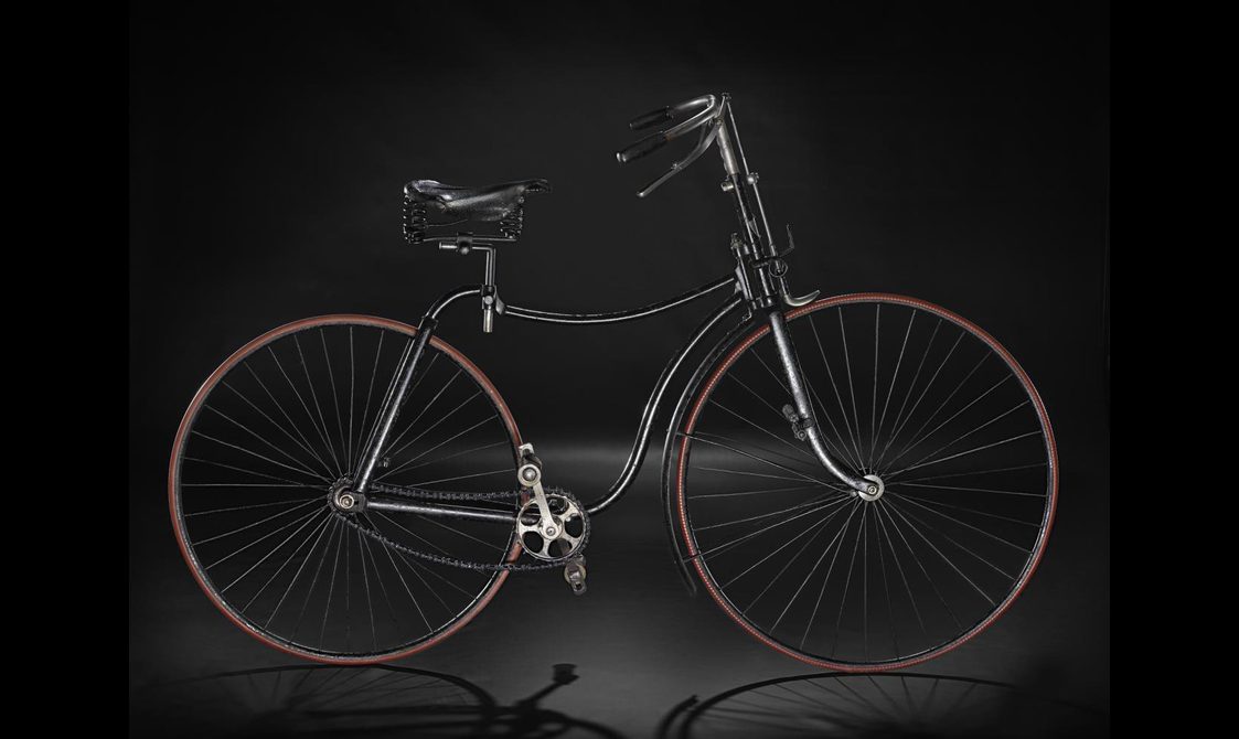 A vintage black bicycle with a light brown rim around the wheels, viewed from the side and standing upright against a black background.