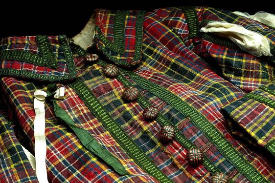 A close up photograph of the royal archers green abd red tartan uniform , with buttons at the front and a green and yellow ribbon on the left side of the jacket. set against black background.