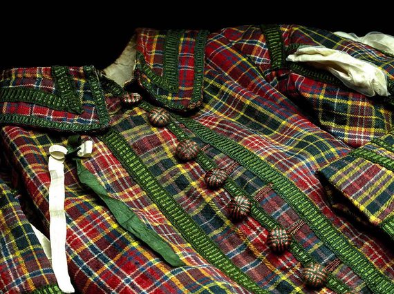A close up photograph of the royal archers green abd red tartan uniform , with buttons at the front and a green and yellow ribbon on the left side of the jacket. set against black background.
